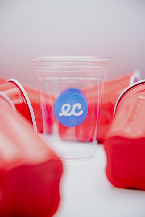 A clear plastic cup with a blue sticker displaying the letters 'ec' is surrounded by several red solo cups. The red cups are haphazardly arranged and provide a stark contrast against the clear cup, which is centrally positioned and highlighted.