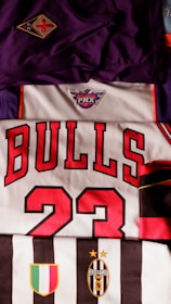 The image features a collection of sports jerseys, including a purple jersey with an emblem, a white jersey with red lettering spelling 'BULLS' and the number 23, and a black and white striped jersey with the Juventus emblem.
