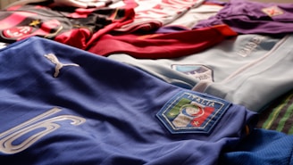 Various football (soccer) jerseys laid out on a surface. The prominently featured jersey in the foreground is blue with the word 'Italia,' a Puma logo, and the number 16. Other jerseys in the background include red, black, white, and purple colors.