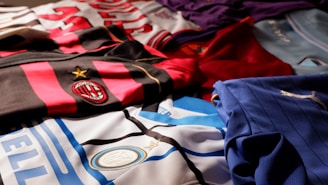 A collection of colorful soccer jerseys is laid out, each featuring distinctive team logos and color patterns. The jerseys include stripes, stars, and various shades, providing a rich visual contrast.