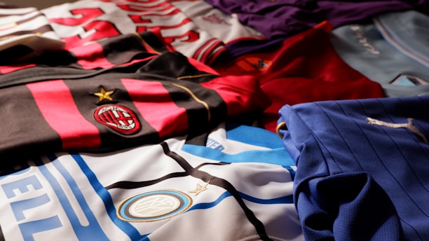 A collection of colorful soccer jerseys is laid out, each featuring distinctive team logos and color patterns. The jerseys include stripes, stars, and various shades, providing a rich visual contrast.