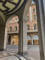 An architectural view through stone arches reveals a street scene with an elegant, ornate building featuring pink and cream colors. Two shops, Missoni and Pollini, have their display windows visible, and a few people are walking by. The design includes intricate window moldings and balustrades on a higher level.