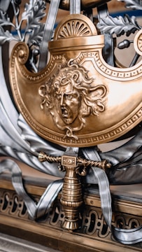A detailed and ornate metallic artwork features a fierce, sculpted face with flowing hair, reminiscent of mythological themes. The design is predominantly in gold and silver tones, incorporating elements like a sword hilt or handle, laurel leaves, and decorative scrollwork.
