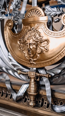 A detailed and ornate metallic artwork features a fierce, sculpted face with flowing hair, reminiscent of mythological themes. The design is predominantly in gold and silver tones, incorporating elements like a sword hilt or handle, laurel leaves, and decorative scrollwork.