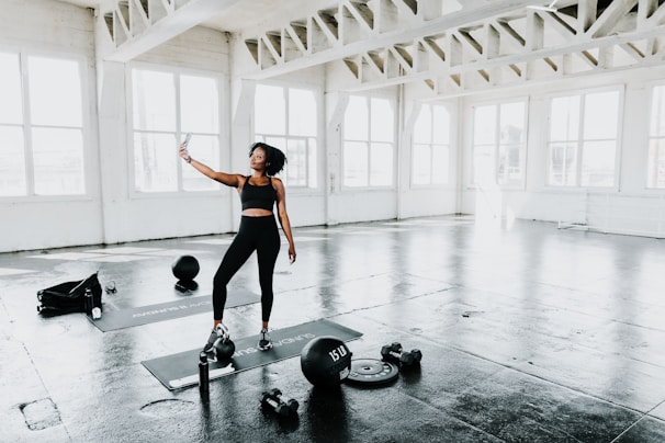 A woman in athletic wear stands in a spacious, well-lit gym with large windows. She holds a smartphone and appears to be taking a selfie. The floor is covered with workout mats and exercise equipment such as kettlebells, weights, and a water bottle are scattered around.
