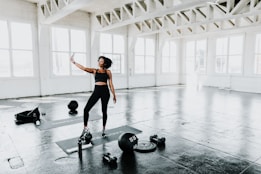 A woman in athletic wear stands in a spacious, well-lit gym with large windows. She holds a smartphone and appears to be taking a selfie. The floor is covered with workout mats and exercise equipment such as kettlebells, weights, and a water bottle are scattered around.