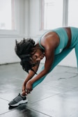A woman in athletic wear is stretching indoors, leaning over to touch her foot on a tiled floor. Natural light streams in through large windows, casting a soft glow over the scene.