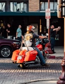 A person wearing a motorcycle helmet and a black vest with a 'Notorious' patch is sitting on a bright orange motorcycle parked on a cobblestone street. The background includes a brick building with large windows and several people, some standing and another taking a photo with a smartphone.
