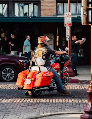 A person wearing a motorcycle helmet and a black vest with a 'Notorious' patch is sitting on a bright orange motorcycle parked on a cobblestone street. The background includes a brick building with large windows and several people, some standing and another taking a photo with a smartphone.