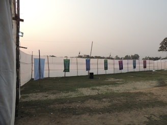 A large outdoor area with a grassy ground and several vertical poles supporting colorful banners. The banners display text and are mounted against a backdrop of a tall white fabric wall stretching across the scene. There is a calm, overcast sky, and a few trees are visible in the distance.