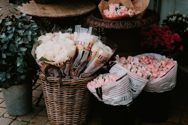 A display of various flowers in a rustic setting, including buckets filled with pink and white roses and tulips. The flowers are organized in wicker baskets and brown paper, surrounded by greenery in a metal container. The arrangement appears to be part of a florists' outdoor setup with cobblestone flooring.