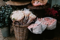 A display of various flowers in a rustic setting, including buckets filled with pink and white roses and tulips. The flowers are organized in wicker baskets and brown paper, surrounded by greenery in a metal container. The arrangement appears to be part of a florists' outdoor setup with cobblestone flooring.