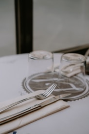 A minimalist table setting featuring a fork and knife neatly placed on a folded napkin. Two glasses are positioned upside down beside the cutlery on a white table with a circular abstract pattern.