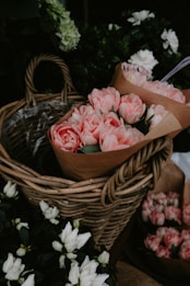 A wicker basket holds a large bouquet of delicate pink flowers, wrapped in brown paper. Surrounding the basket are other floral arrangements featuring white and pink blossoms. The greenery provides a lush backdrop, enhancing the vivid colors of the flowers.