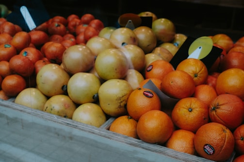 A variety of fruits are arranged in a market display. The image features a selection of oranges and grapefruits placed neatly in wooden crates. Some of the fruits have stickers on them, indicating they are for sale. The overall composition is orderly and colorful, with the different shades of orange and yellow creating a warm visual appeal.
