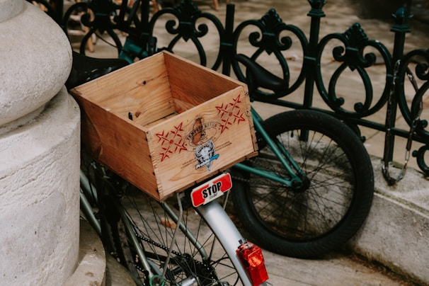 A wooden crate is attached to the back of a bicycle, with a decorative and ornate black iron fence in the background. The crate features some engraved graphics. The rear wheel of the bicycle is visible, along with a small stop sign attached near the rear light. The surroundings include a stone column and pavement.
