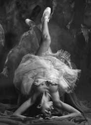 A ballet dancer is captured in an elegant pose, balancing upside-down with legs gracefully extended. The dancer's tutu creates a dynamic and flowing shape, contrasting with the dark, textured background. The image is in black and white, emphasizing the play of light and shadow across the dancer's form.