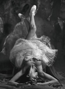 A ballet dancer is captured in an elegant pose, balancing upside-down with legs gracefully extended. The dancer's tutu creates a dynamic and flowing shape, contrasting with the dark, textured background. The image is in black and white, emphasizing the play of light and shadow across the dancer's form.