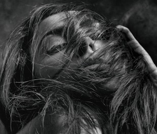 A close-up black and white photograph of a person with long hair partially covering their face. The individual has their eyes closed and their hand near their hair, giving the impression of being lost in thought or emotion.