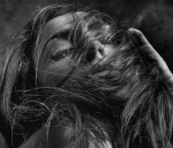 A close-up black and white photograph of a person with long hair partially covering their face. The individual has their eyes closed and their hand near their hair, giving the impression of being lost in thought or emotion.