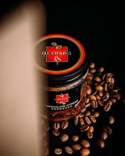 A jar of chocolate spread with an espresso flavor is placed on a black surface, surrounded by scattered coffee beans. The lid of the jar prominently displays a logo with the words 'Espoleto'. A soft light casts a shadow on the left side, creating a dramatic effect.