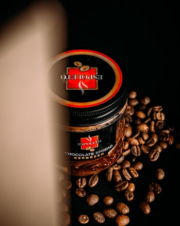 A jar of chocolate spread with an espresso flavor is placed on a black surface, surrounded by scattered coffee beans. The lid of the jar prominently displays a logo with the words 'Espoleto'. A soft light casts a shadow on the left side, creating a dramatic effect.