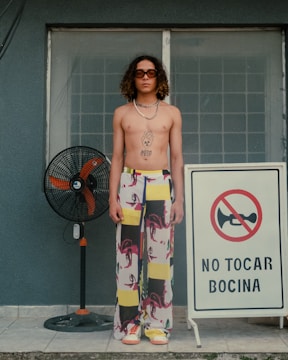 A person with curly hair and sunglasses stands shirtless in front of a barred window. They wear brightly patterned pants and sneakers. Next to them is a standing fan with orange blades, and a sign with a crossed-out horn symbol and the text 'NO TOCAR BOCINA.'