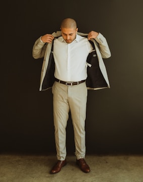 A man stands against a dark background, adjusting his light-colored suit jacket with a white shirt underneath. He is wearing brown dress shoes and appears to be getting ready or straightening his attire.