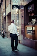 A man in formal attire is walking along a city street, passing by storefronts with prominent signage including Omega and Cartel. The scene suggests a business district atmosphere with modern architectural elements.