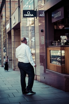 A man in formal attire is walking along a city street, passing by storefronts with prominent signage including Omega and Cartel. The scene suggests a business district atmosphere with modern architectural elements.