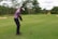 A golfer in a purple floral shirt and dark pants is captured mid-swing on a lush green golf course. The surroundings include well-maintained grass, several sand traps, and tall trees lining the course. The sky is overcast, adding a calm atmosphere to the scene.