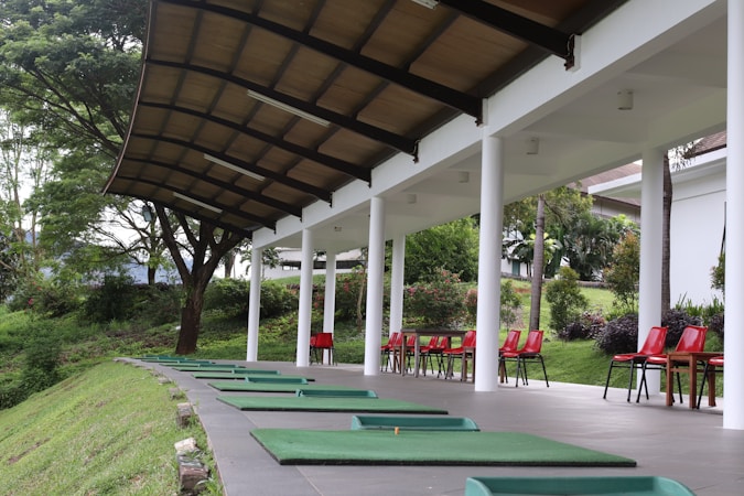 A driving range with green mats arranged in a row under a covered structure. Red chairs are placed next to each mat. The surrounding area has lush greenery and trees.