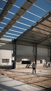 An industrial construction site with visible steel framework and a partially completed roof under a clear blue sky. Two workers are present, one standing on the ground observing the structure, and another perched on the framework above. The ground is covered in dirt and construction materials.