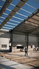 An industrial construction site with visible steel framework and a partially completed roof under a clear blue sky. Two workers are present, one standing on the ground observing the structure, and another perched on the framework above. The ground is covered in dirt and construction materials.