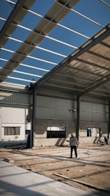 An industrial construction site with visible steel framework and a partially completed roof under a clear blue sky. Two workers are present, one standing on the ground observing the structure, and another perched on the framework above. The ground is covered in dirt and construction materials.