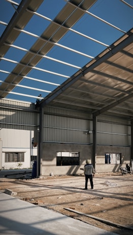 An industrial construction site with visible steel framework and a partially completed roof under a clear blue sky. Two workers are present, one standing on the ground observing the structure, and another perched on the framework above. The ground is covered in dirt and construction materials.