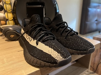 A pair of black athletic shoes with a distinctive white stripe and text on the side is placed on a wooden surface. Nearby, a set of black over-ear headphones and a coffee pod holder are visible, suggesting a casual and modern setting.