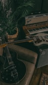 A cozy corner filled with music equipment, including an electric guitar resting on a brown sofa, an amplifier with the Fender logo, a vintage keyboard, and a woven basket containing a lush green plant. There's a sense of a creative, relaxed atmosphere.