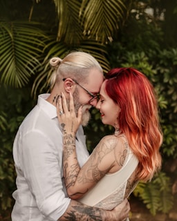 A couple lovingly embraces, touching foreheads while smiling warmly at each other. The man has a beard and wears glasses, with his hair tied back. The woman has long, red hair and both display intricate tattoos on their arms. They are surrounded by lush, green foliage, creating an intimate setting.