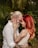 A couple lovingly embraces, touching foreheads while smiling warmly at each other. The man has a beard and wears glasses, with his hair tied back. The woman has long, red hair and both display intricate tattoos on their arms. They are surrounded by lush, green foliage, creating an intimate setting.