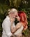 A couple lovingly embraces, touching foreheads while smiling warmly at each other. The man has a beard and wears glasses, with his hair tied back. The woman has long, red hair and both display intricate tattoos on their arms. They are surrounded by lush, green foliage, creating an intimate setting.
