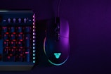 A black gaming mouse and a section of a mechanical keyboard are illuminated by neon lights against a dark backdrop. The mouse is displaying a glowing logo, and the keyboard has prominently lit keys with numbers on them. The lighting creates a vibrant contrast between the deep purple and bluish hues.