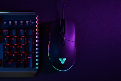 A black gaming mouse and a section of a mechanical keyboard are illuminated by neon lights against a dark backdrop. The mouse is displaying a glowing logo, and the keyboard has prominently lit keys with numbers on them. The lighting creates a vibrant contrast between the deep purple and bluish hues.