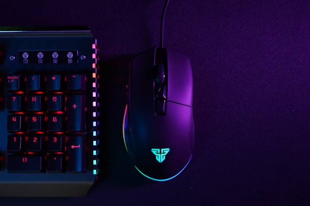 A black gaming mouse and a section of a mechanical keyboard are illuminated by neon lights against a dark backdrop. The mouse is displaying a glowing logo, and the keyboard has prominently lit keys with numbers on them. The lighting creates a vibrant contrast between the deep purple and bluish hues.