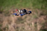 A drone with blue propellers and a mounted camera is flying in mid-air against a blurred grassy background.