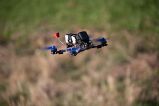 A drone with blue propellers and a mounted camera is flying in mid-air against a blurred grassy background.