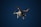 A white drone with four rotors is flying in the sky. The drone is visible against a dark blue sky and appears to have a camera mounted at its base. The rotors are in motion, and there are red lights on the drone's body.