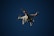 A white drone with four rotors is flying in the sky. The drone is visible against a dark blue sky and appears to have a camera mounted at its base. The rotors are in motion, and there are red lights on the drone's body.