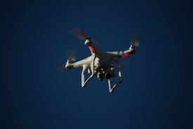 A white drone with four rotors is flying in the sky. The drone is visible against a dark blue sky and appears to have a camera mounted at its base. The rotors are in motion, and there are red lights on the drone's body.