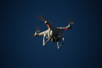 A white drone with four rotors is flying in the sky. The drone is visible against a dark blue sky and appears to have a camera mounted at its base. The rotors are in motion, and there are red lights on the drone's body.